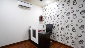 A room with a wooden floor, a black office chair, a white desk with a red object on it, and a wall with a circular pattern. - Hotel Mannat International (New Delhi)