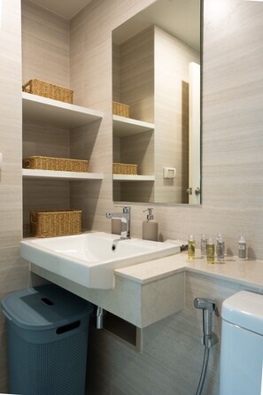 Un baño moderno con un lavamanos grande, espejo, cestas de mimbre en estantes, artículos de aseo en la encimera y un cubo de basura.
