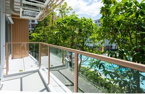 Un balcón moderno al aire libre con barandillas de vidrio, un piso de madera y vista a una piscina rodeada de abundante vegetación y edificios.