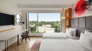 A modern hotel room with a large window, a flat-screen TV, a desk, and two beds.