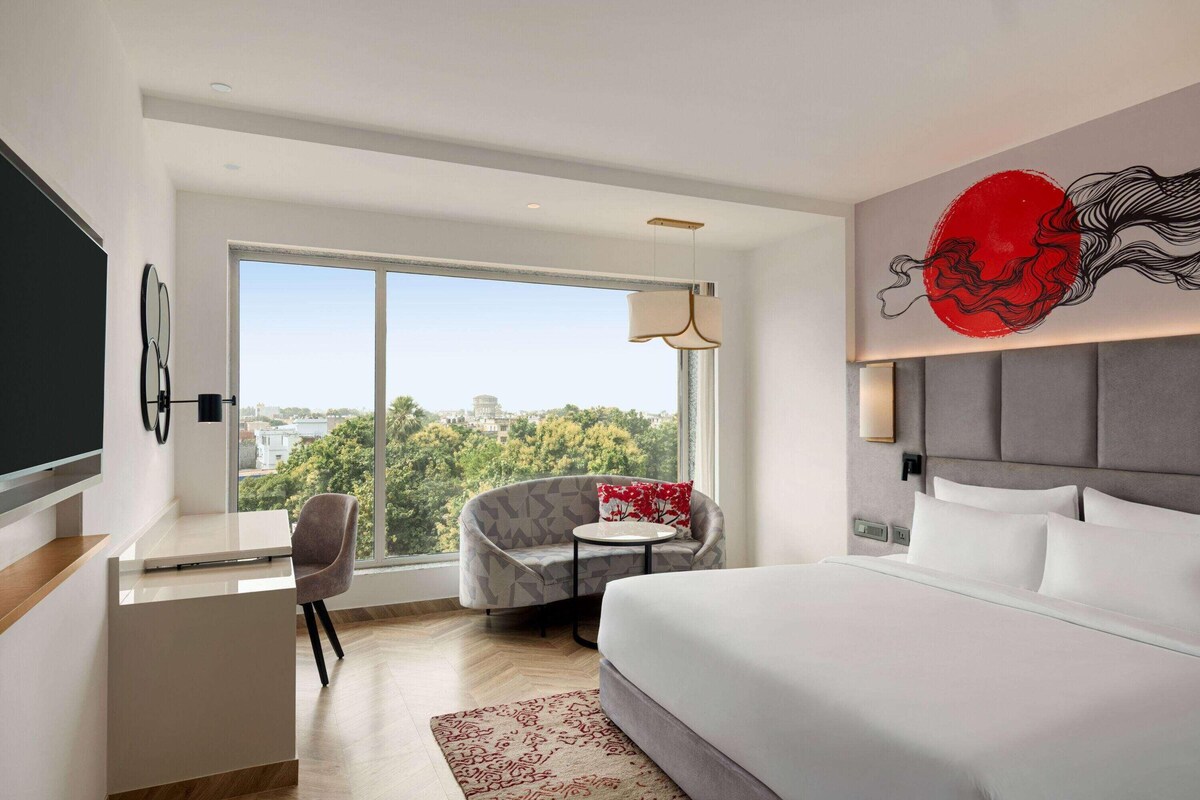 A modern hotel room with a large bed, a desk, a chair, and a sofa. There is a large window with a view of the city.