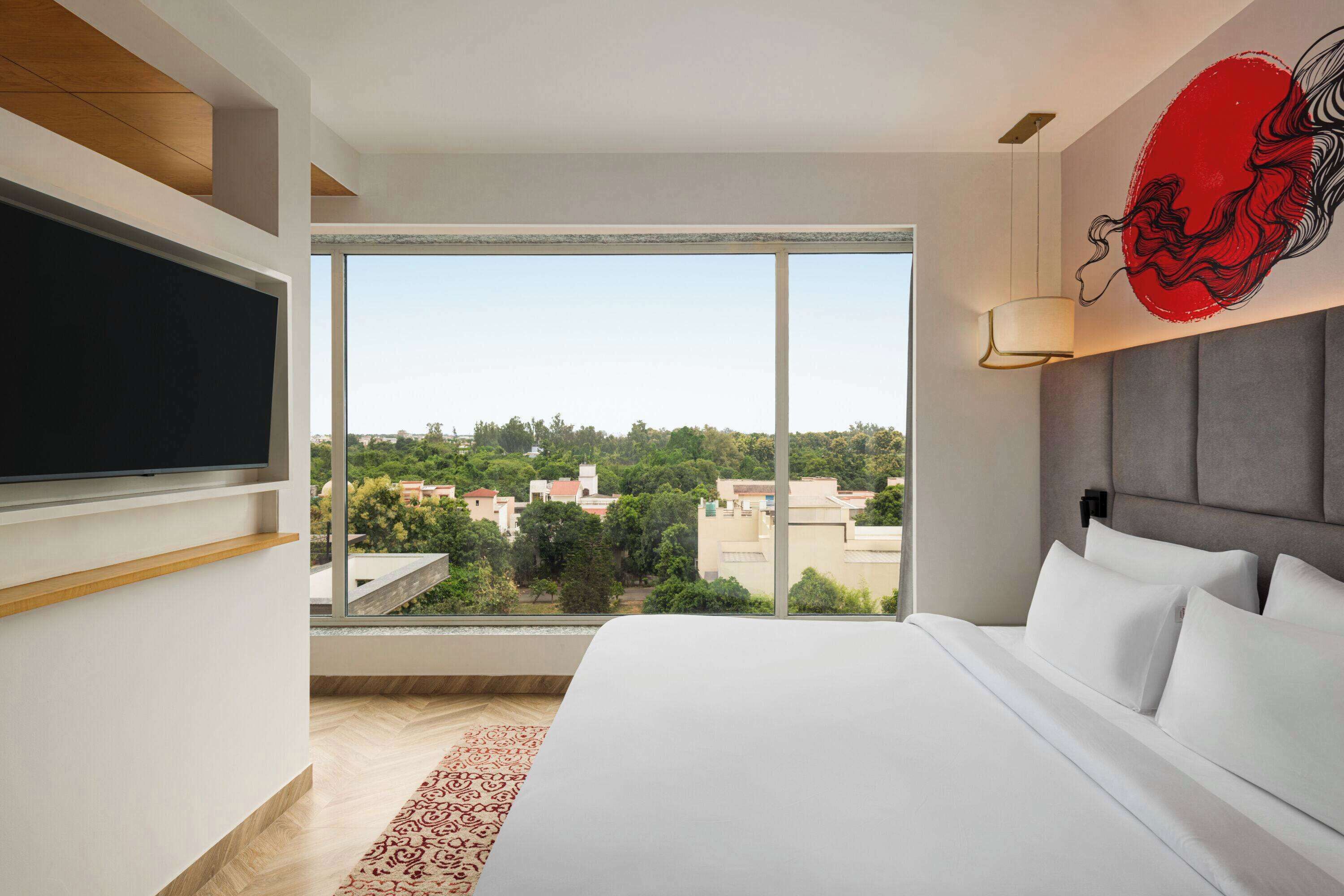 Ein modernes Hotelzimmer mit einem großen Bett, einem flachbildfernseher an der Wand und einem großen Fenster mit Blick auf die Stadt.