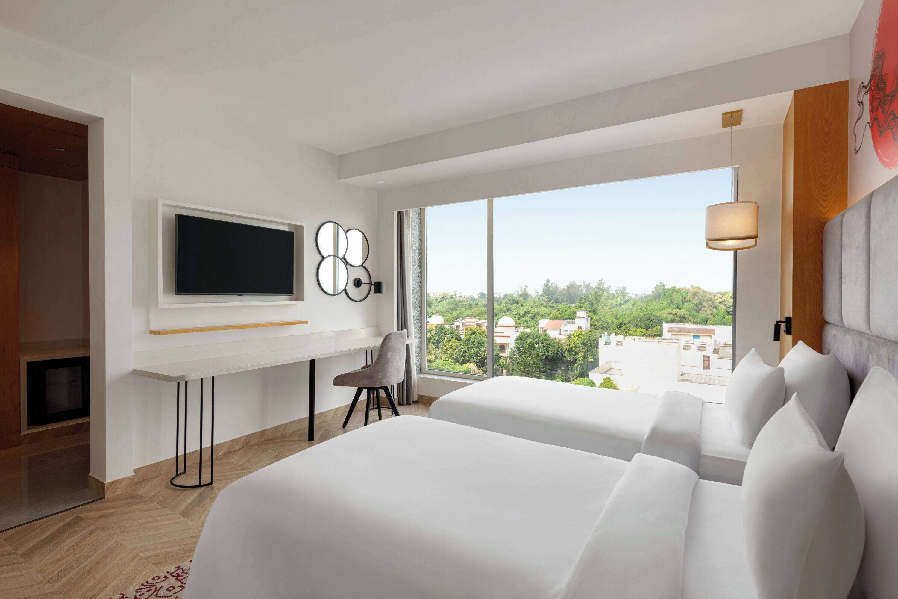 A modern hotel room with a large bed, a desk with a flat-screen TV, a chair, and a large window offering a city view.