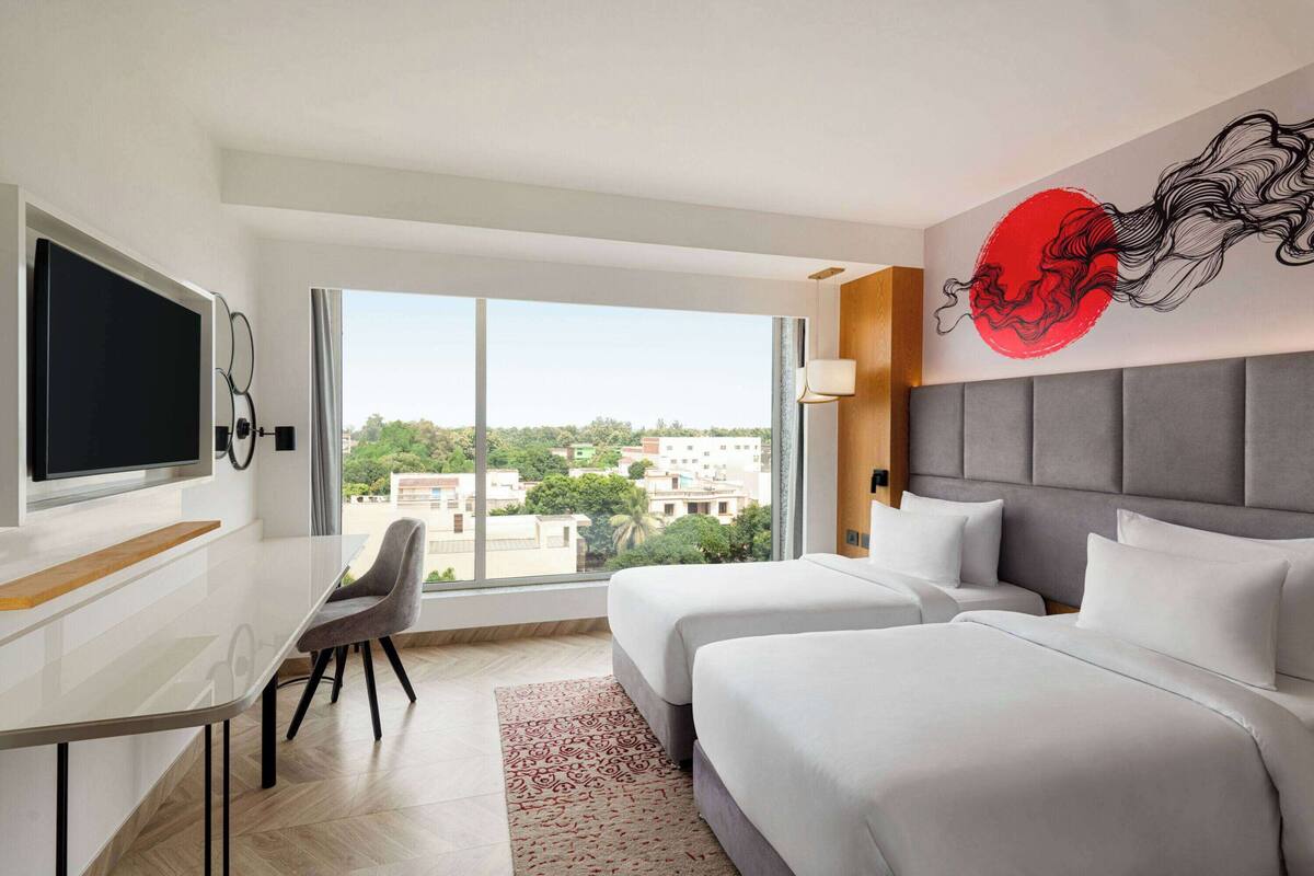 Ein Hotelzimmer mit zwei Betten, einem Schreibtisch, einem Fernseher und einem großen Fenster mit Blick auf die Stadt.
