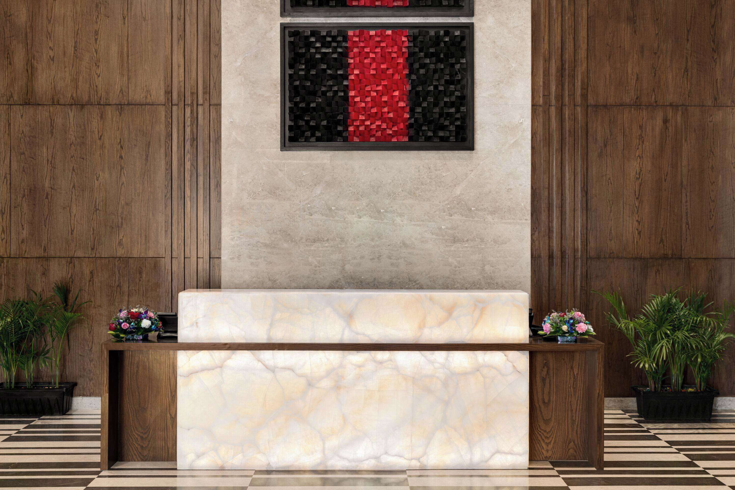 Balcão de recepção com bancada de mármore, ladeado por duas plantas em vasos, e um painel na parede com padrão em vermelho e preto.