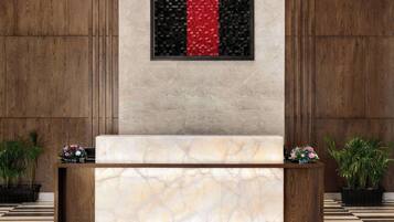 Balcão de recepção com bancada de mármore, ladeado por duas plantas em vasos, e um painel na parede com padrão em vermelho e preto.