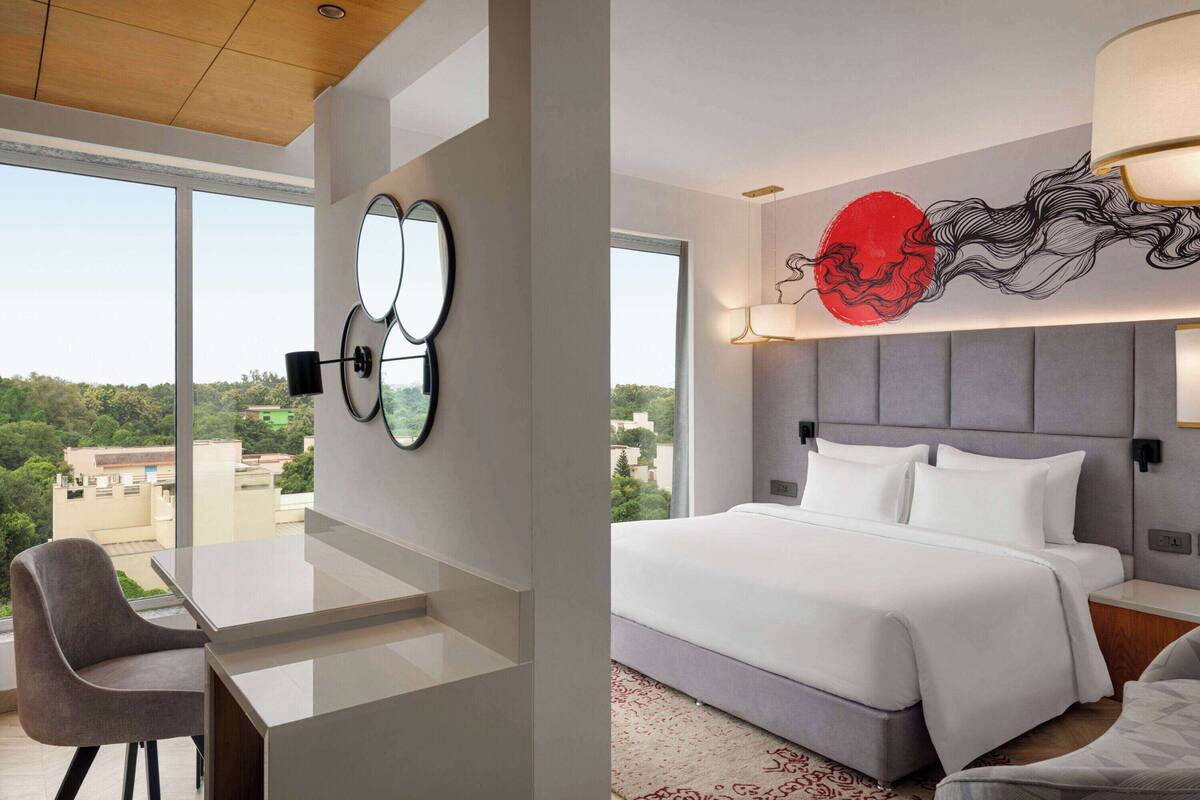 Ein modernes Hotelzimmer mit einem großen Bett, einem Schreibtisch und einem Stuhl. Ein großes Fenster bietet Blick auf die Stadt.