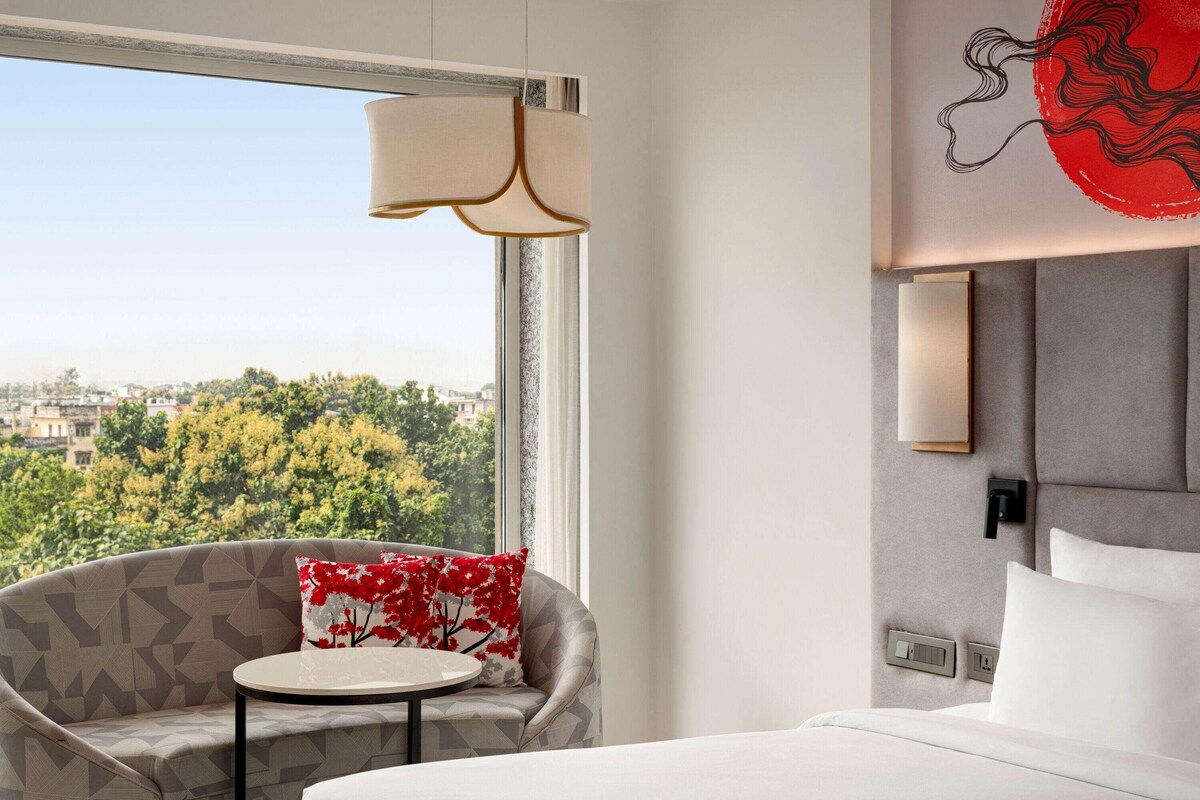 Quarto de hotel com vista para árvores e edifícios, um sofá com almofada vermelha, uma mesa redonda e uma cama com lençóis brancos.