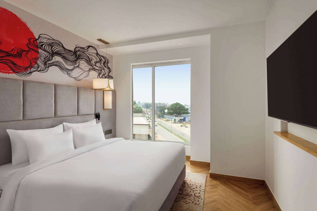 Ein modernes Hotelzimmer mit einem großen Bett, Blick auf die Stadt durch eine Schiebetür und einem an der Wand befestigten Flachbildfernseher.