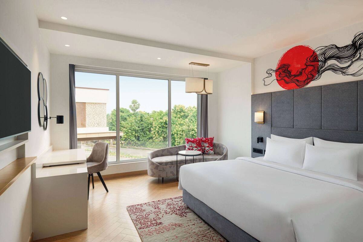 Ein modernes Hotelzimmer mit einem großen Bett, einem Schreibtisch, einem Sessel und Blick auf Grünanlagen durch das Fenster.