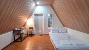 Un dormitorio de buhardilla pequeño y bien iluminado, con techo inclinado de madera, una cama individual con colcha estampada, un escritorio pequeño con silla y una ventana con vistas al exterior.