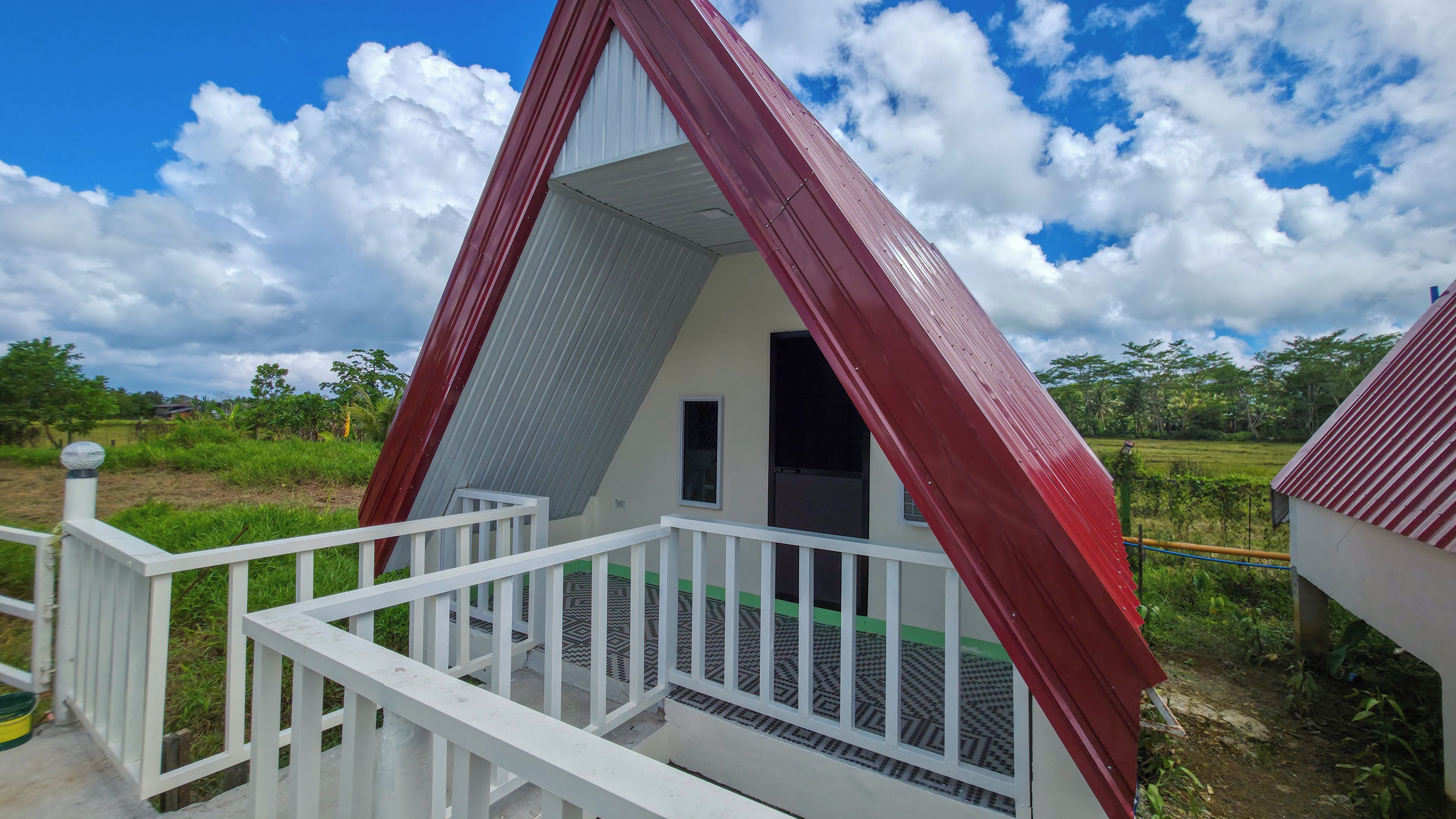 Um pequeno edifício triangular com telhado vermelho e paredes brancas, apresentando uma varanda com grade branca.