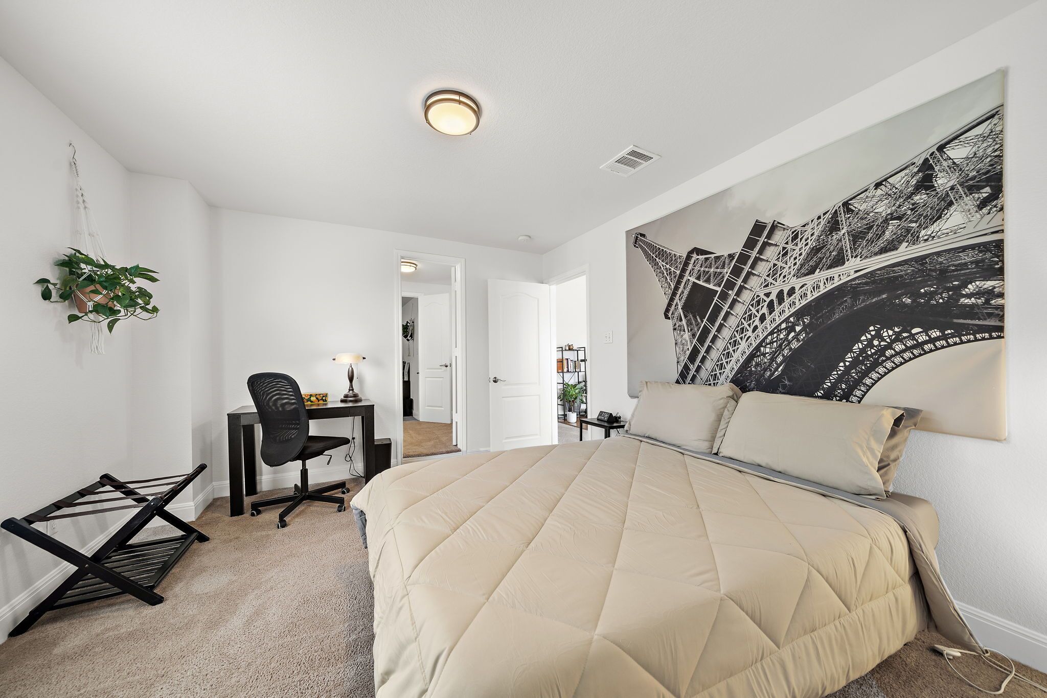 Ein Schlafzimmer mit einem großen Bett, einem Schreibtisch mit Stuhl, einer hängenden Pflanze und einem Bild des Eiffelturms.
