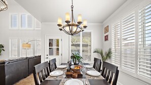 Uma sala de jantar com uma mesa de madeira escura posta para seis pessoas, cadeiras brancas, um candelabro e vista para o exterior através de persianas brancas.
