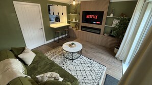Sala de estar com sofá verde, mesa de centro branca, TV fixada em uma parede de madeira e uma área de cozinha ao fundo.