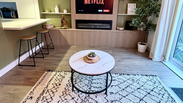 Uma sala de estar moderna com uma TV de tela plana exibindo 'NETFLIX', uma estante de madeira com prateleiras, uma mesa de centro redonda com uma planta e um lustre.