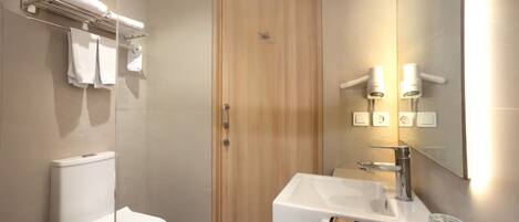 Un baño moderno con inodoro blanco, puerta de madera, lavamanos con espejo y toallero con toallas dobladas.