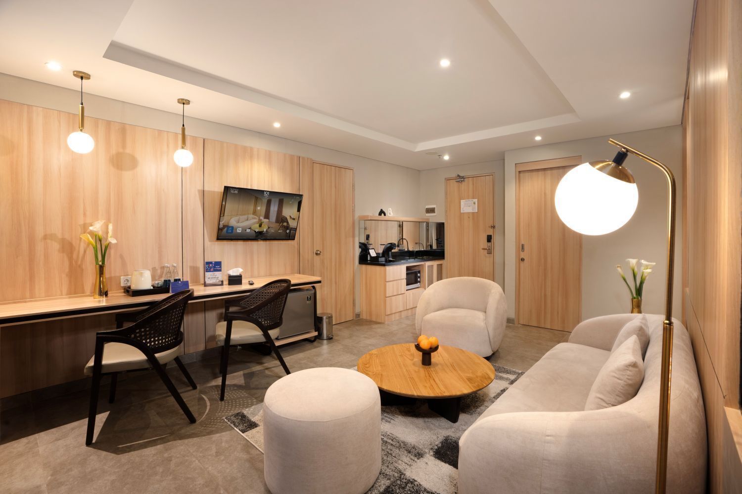 A modern hotel room with a wooden accent wall, a flat-screen TV, a kitchenette, a sofa, a round coffee table, and a desk with a chair.