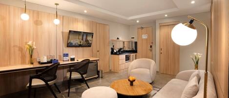 A modern hotel room with a wooden accent wall, a flat-screen TV, a kitchenette, a sofa, a round coffee table, and a desk with a chair.