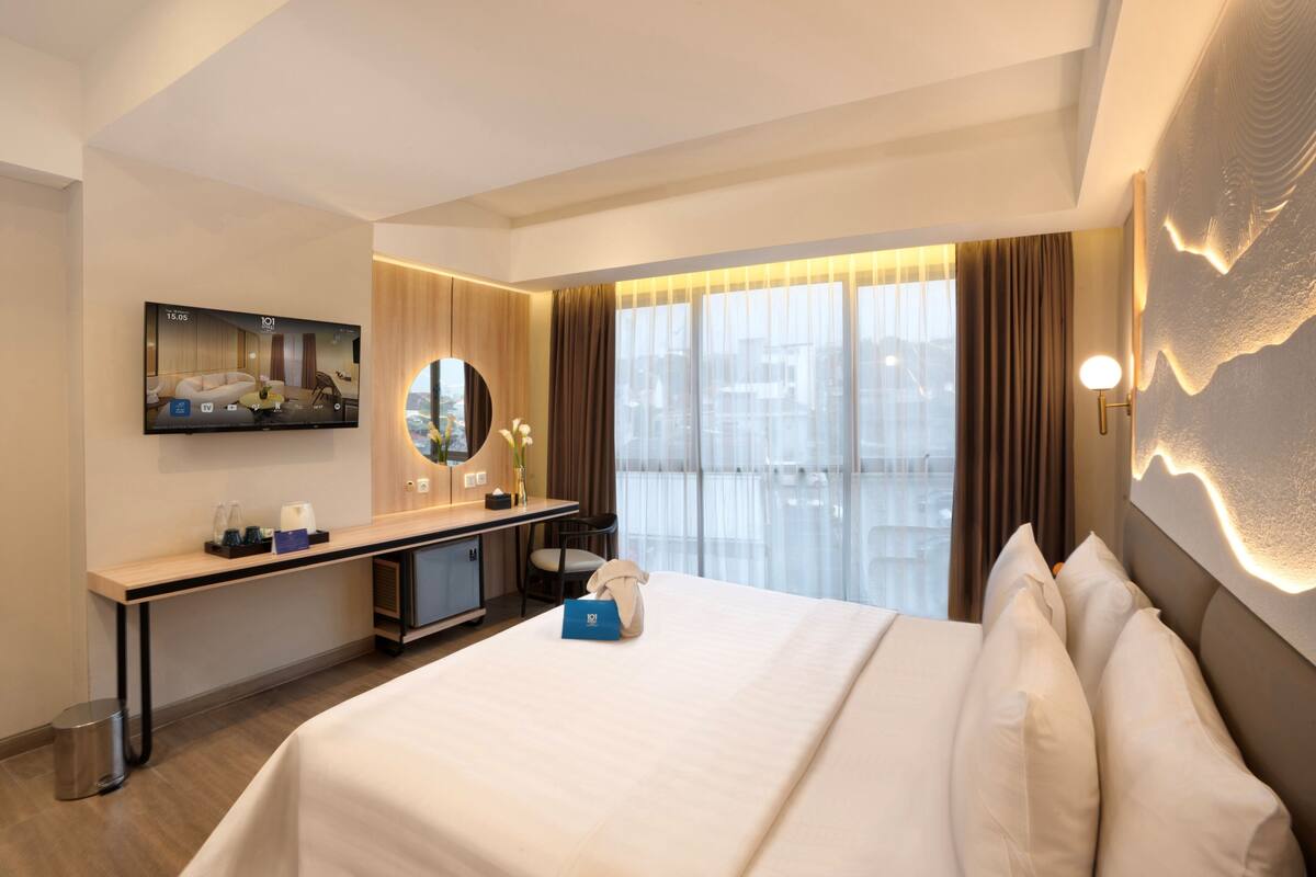 A hotel room with a bed, a desk with a computer, a TV, and a large window with curtains.