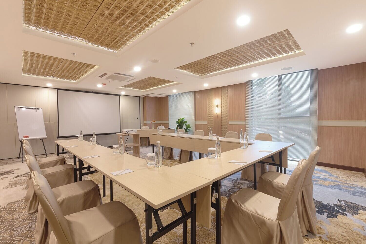 A conference room with a long table, beige chairs, a whiteboard, and a projector screen.