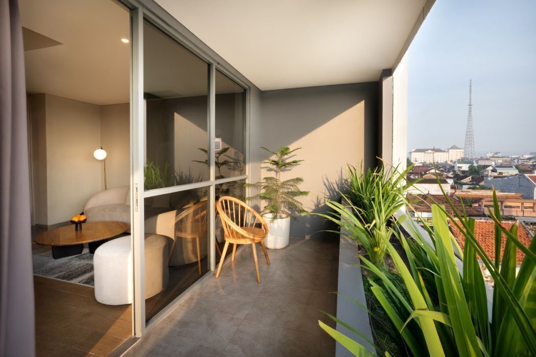 A modern balcony with a glass door, a sofa, a wooden chair, and potted plants.