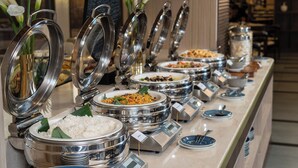 Un bufé con diversas opciones de comida, incluyendo arroz, fideos y otros platos, presentados en una barra con recipientes metálicos.