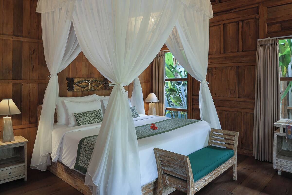 A wooden room with a four-poster bed, a nightstand with a lamp, and a window with curtains.