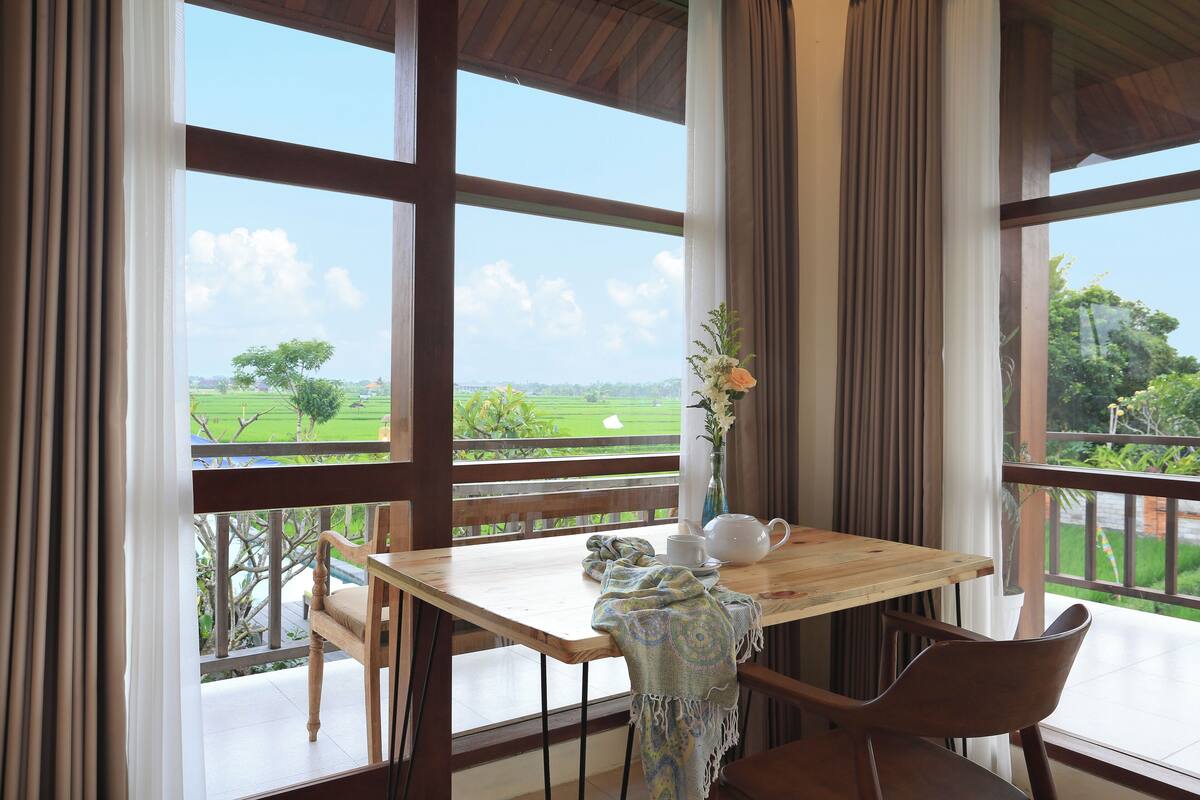 A wooden table with a vase of flowers, a teapot, and a cup on a balcony with a view of a green field.