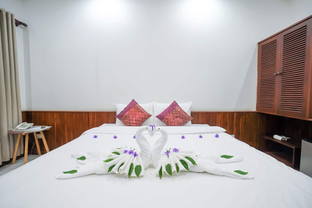 Habitación de hotel con una cama bien hecha, dos almohadones decorativos, un armario de madera y una mesita con un teléfono.