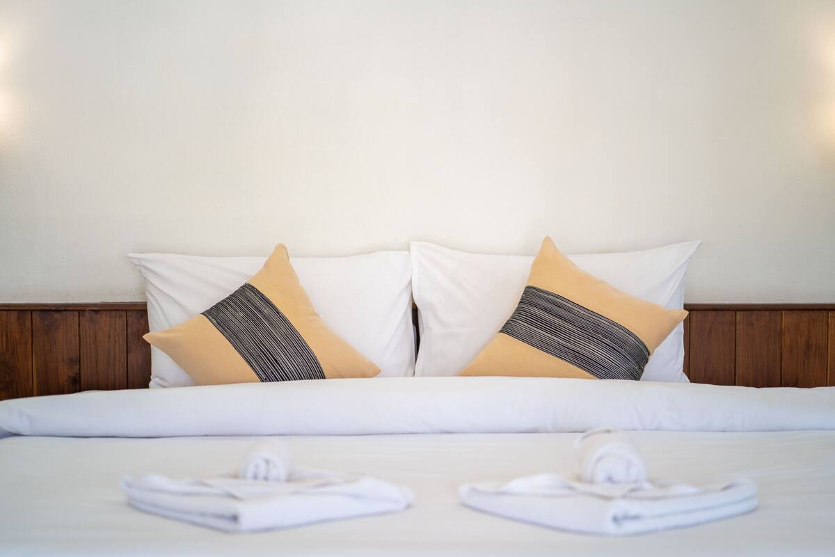 Una cama bien hecha con sábanas blancas y dos almohadas decorativas, una beige y otra con estampado blanco y negro.