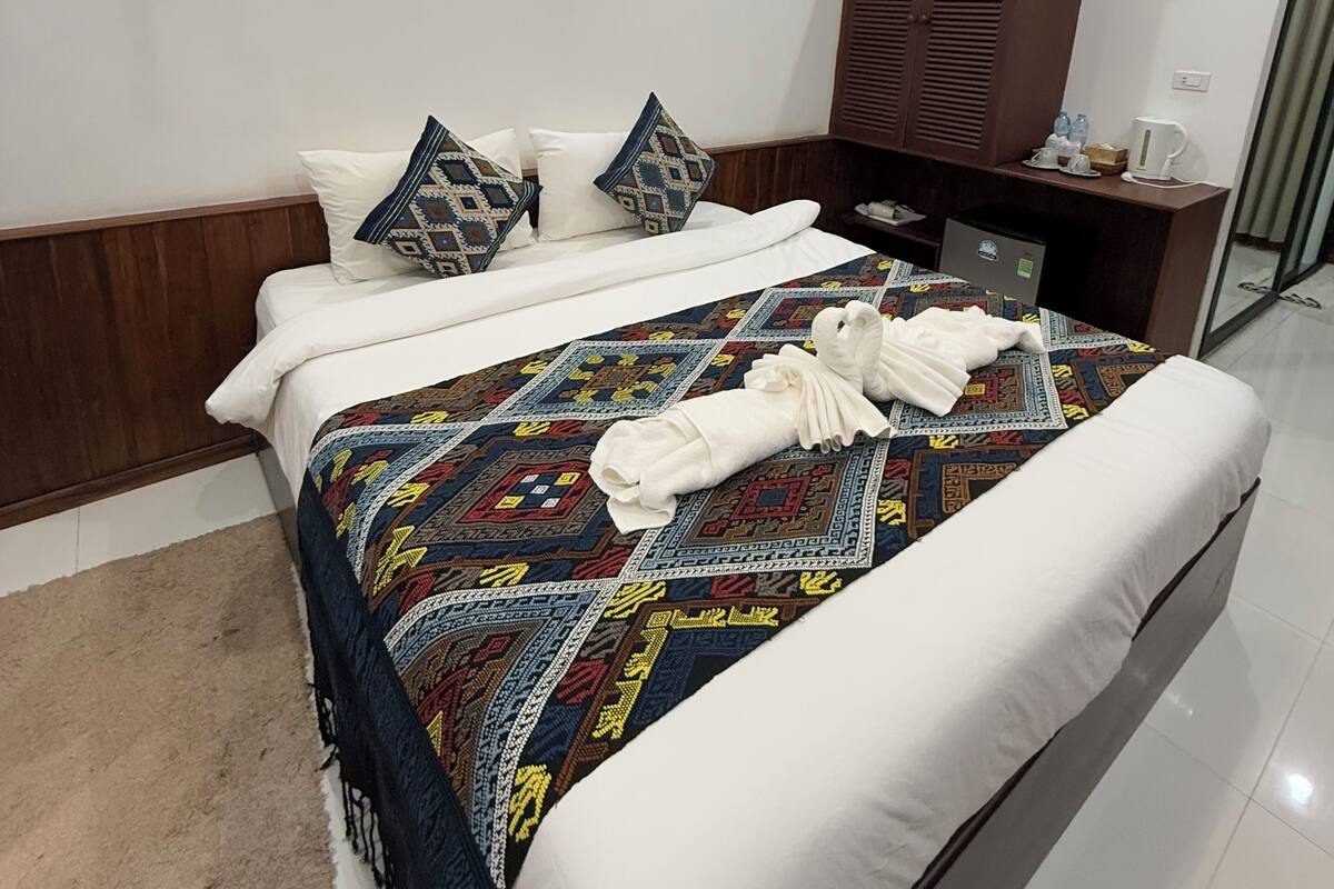 Una cama bien hecha con una colcha estampada y almohadas, un cabecero de madera, un armario y una mesita de noche con hervidor y botellas de agua.