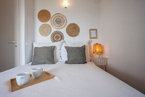 Un lit bien fait, avec une literie blanche et des coussins à motifs noir et blanc, un plateau en bois sur lequel reposent deux tasses blanches, et un mur décoré de tentures murales tissées et d’un tableau encadré.