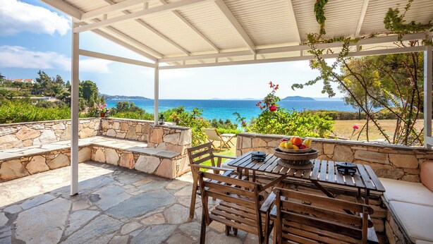 Ein überdachter Außenbereich mit Steinwänden, einem Tisch mit Stühlen und Blick auf das Meer.