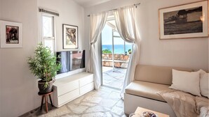 Ein Wohnzimmer mit Sofa, einer Topfpflanze, einem Fernseher und Meerblick durch ein Fenster.