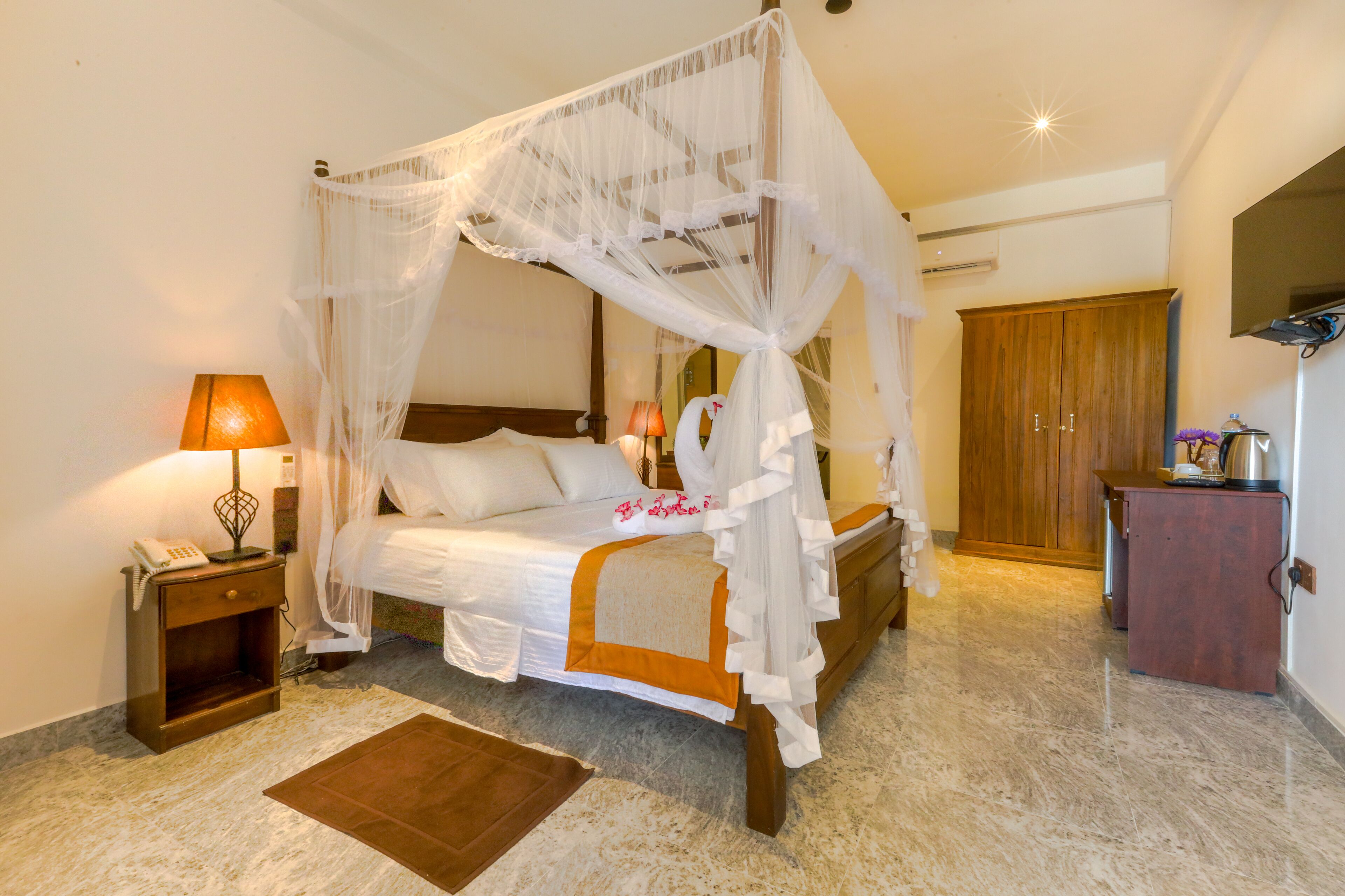 A four-poster bed with a canopy, a bedside table with a lamp, a wooden wardrobe, and a small cabinet with a kettle.
