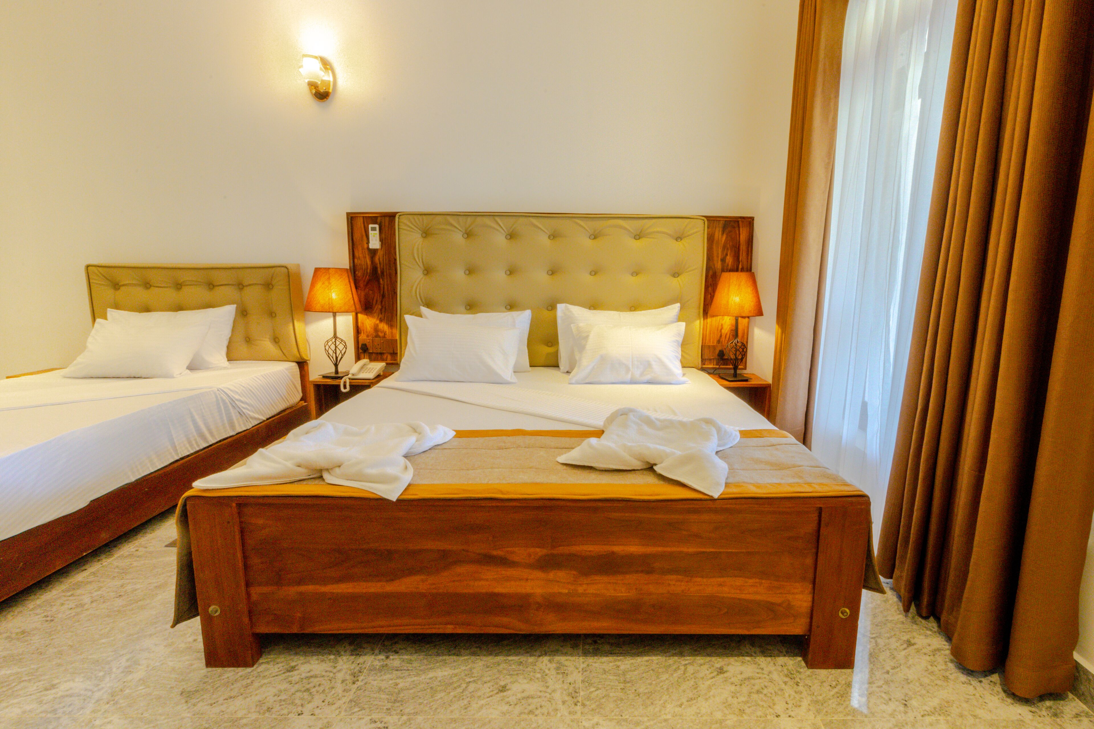 A hotel room with two single beds and a larger bed, all with white linens. There are two bedside lamps and a wall-mounted light. The room has a wooden headboard and a large window with curtains.