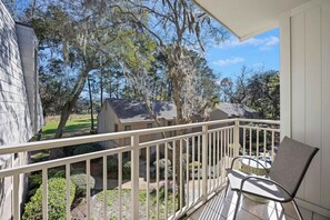 A balcony with a chair, a view of a residential area with trees and houses, and a clear sky.