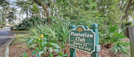 Une enseigne indiquant « Plantation Club », avec le chiffre 10, entourée d’une végétation luxuriante et avec une route en arrière-plan.