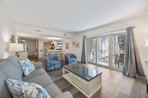 A modern living room with a grey sofa, blue patterned armchairs, a glass coffee table, and a view through large sliding glass doors to an outdoor balcony.