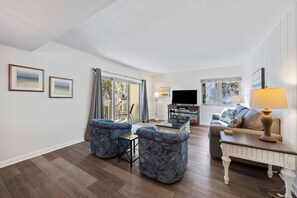 Un salon comprenant un canapé, deux fauteuils, une table basse, un téléviseur et une fenêtre avec des rideaux.