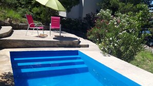 Una piscina con escalones de acceso, rodeada por un jardín con diversas plantas y dos sillas rojas bajo un parasol verde.