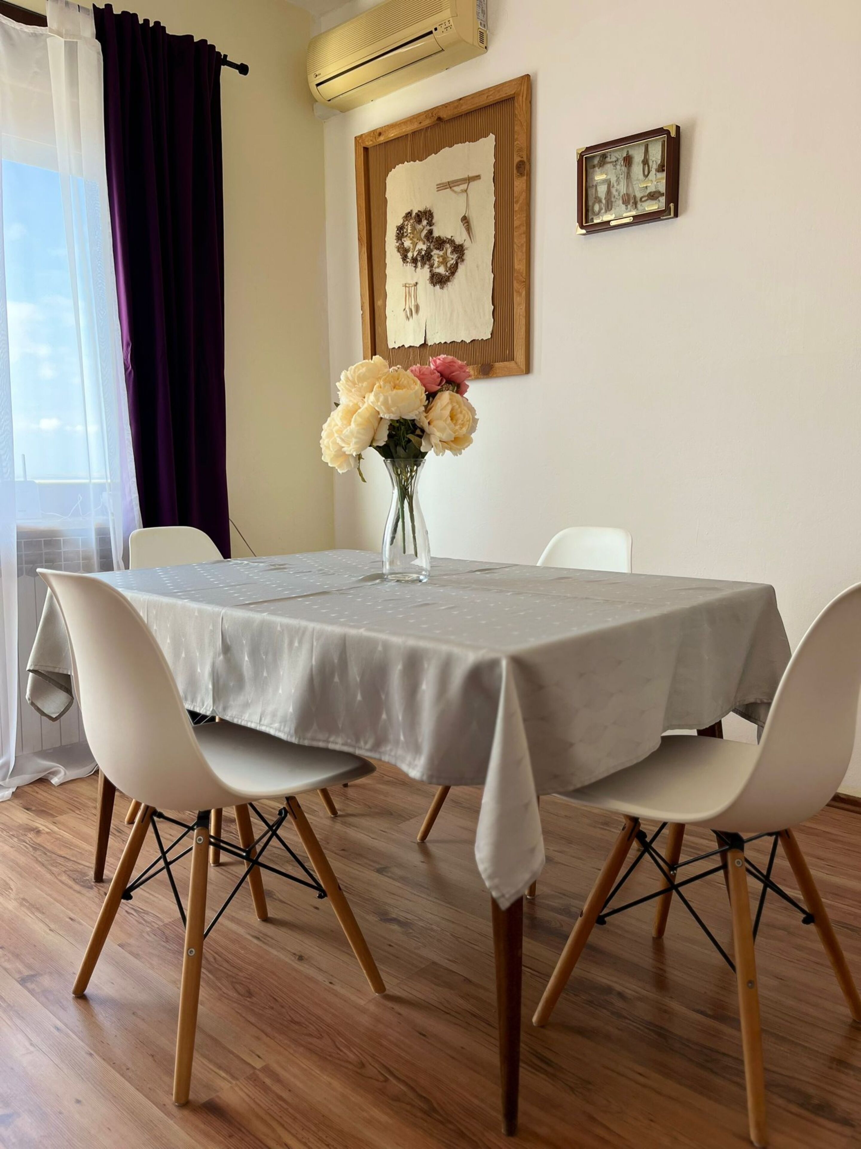 A dining area with a table covered by a grey tablecloth, surrounded by white chairs with wooden legs. A vase with flowers is placed on the table.