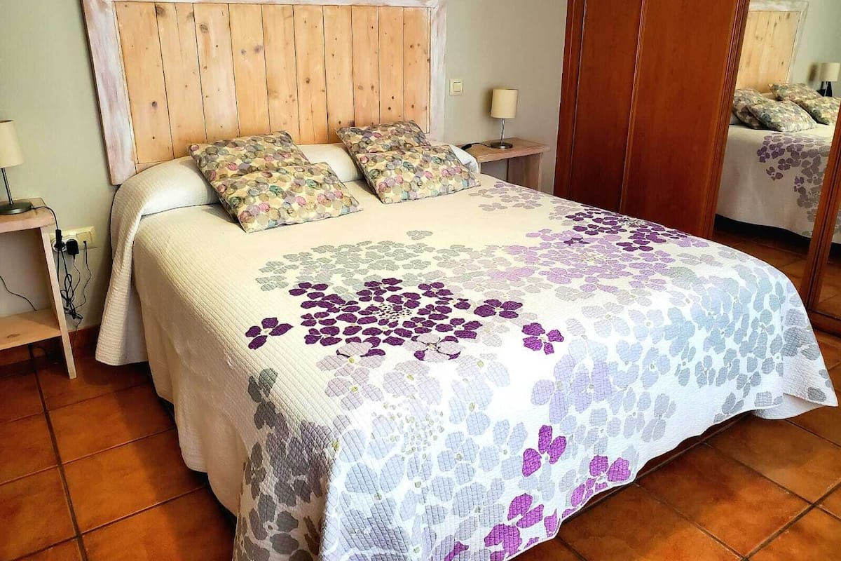 Une chambre à coucher avec une tête de lit en bois, une couvre-lit à motifs floraux, deux coussins assortis, une table de chevet avec une lampe et un miroir au mur.