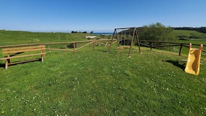 Een gazon met een speelplaats, een schommel en een glijbaan, omgeven door groene velden en een heldere lucht.