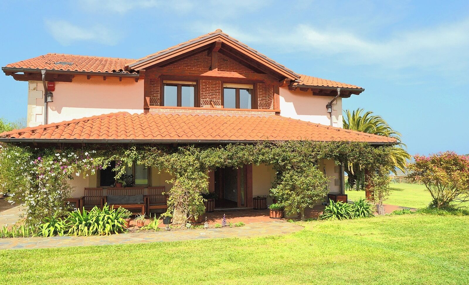 Una casa de dos pisos con techo de tejas, un porche con banco y un jardín cuidado.
