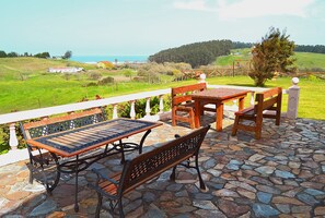 Une terrasse avec un sol en pierre, des meubles en bois et une balustrade blanche donnant sur un paysage verdoyant et la mer.
