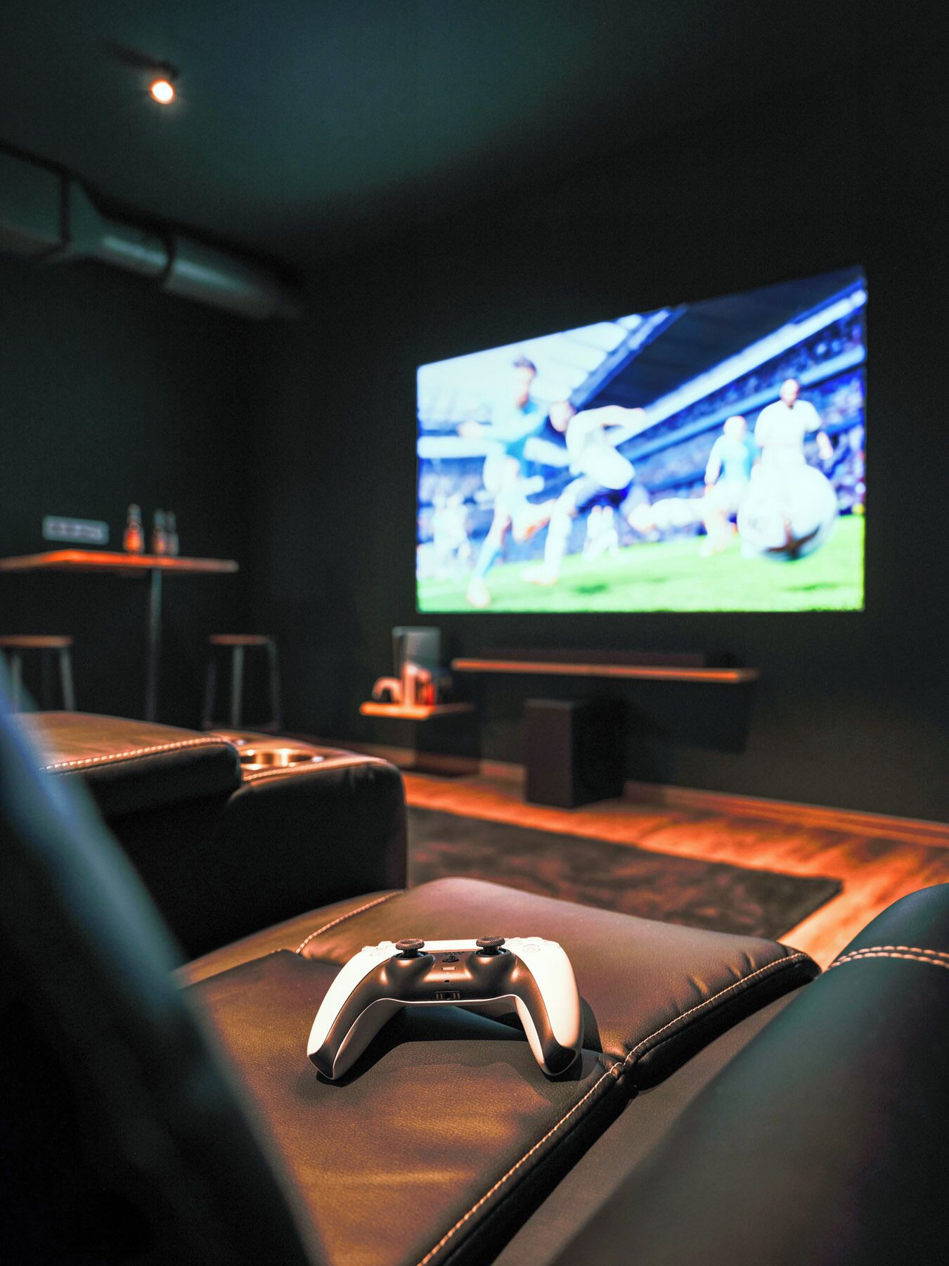 A dark room with a large flat-screen TV displaying a sports event, a leather couch, and a gaming controller on the couch.
