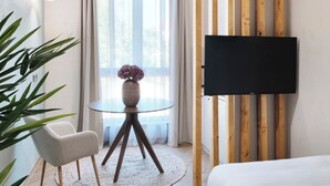 A modern hotel room with a round table, a sofa, a TV mounted on a wooden panel, and a large window with sheer curtains. - PHIL & MAX Hotel & Apartments Messe (Laatzen)