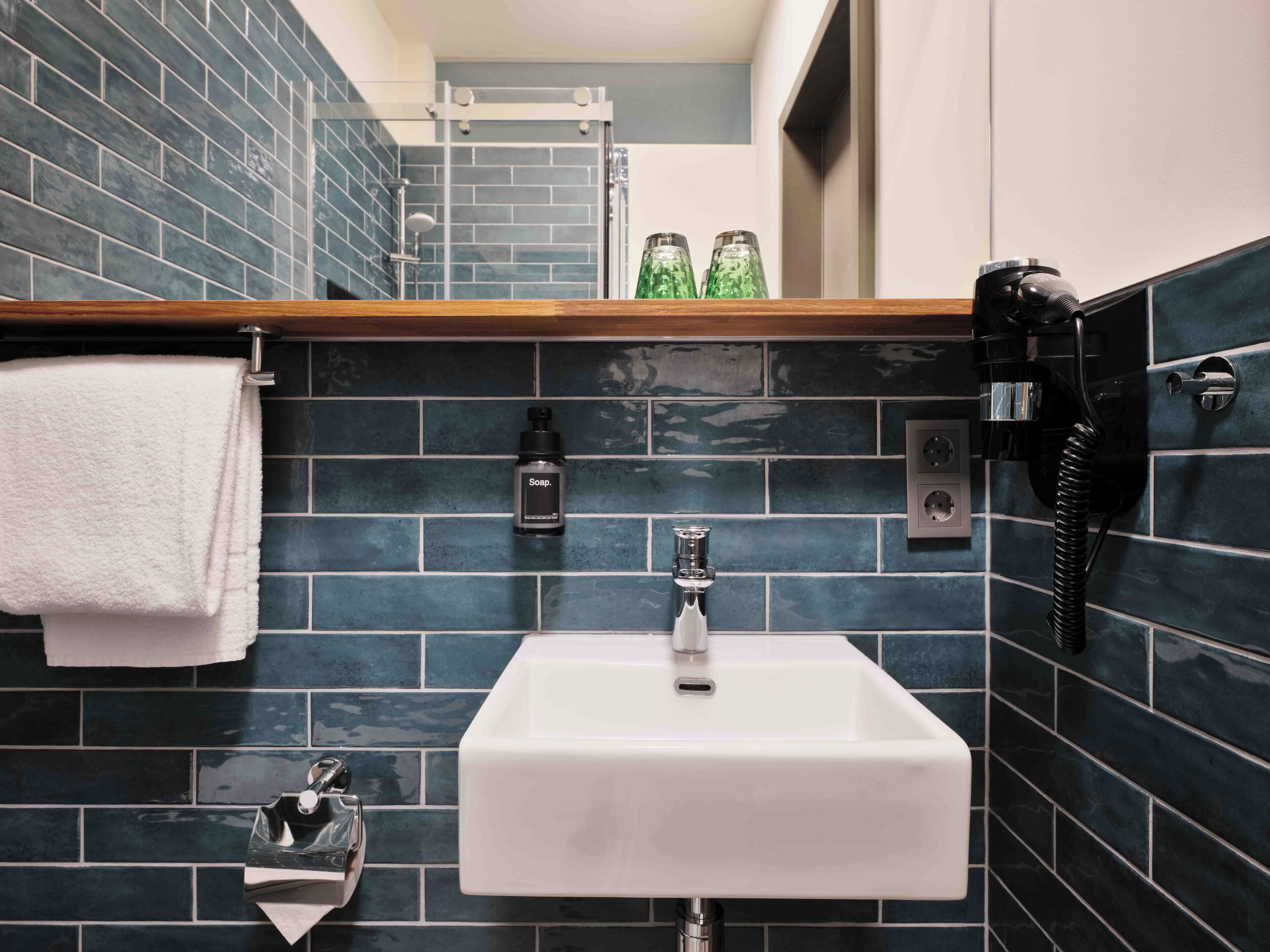 Une salle de bain avec un lavabo blanc, des murs recouverts de carreaux bleu foncé, un plan de toilette en bois, un porte-serviettes, un distributeur de savon et une douchette à main.